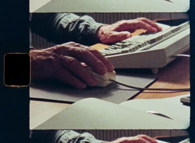 An archival film-style close-up shows a person's hands typing on a vintage computer keyboard and using an old mouse