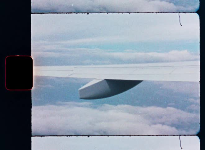 Vintage 8mm film strip footage captures an airplane wing soaring through the sky above the clouds and landscape below