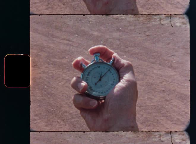 A close-up shot of a person's hand holding a ticking analog stopwatch on vintage 8mm film