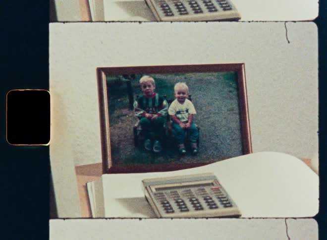 A vintage 8mm film scan reveals a framed photograph of two young brothers next to an old calculator