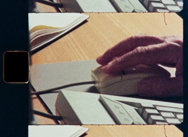 A close-up shot of a hand using a vintage computer mouse on a wooden desk, with an archival film look