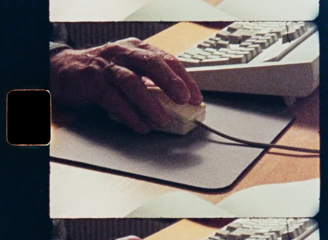 Vintage 8mm film captures an elderly person's hands using a retro computer mouse and typing on a keyboard at a desk