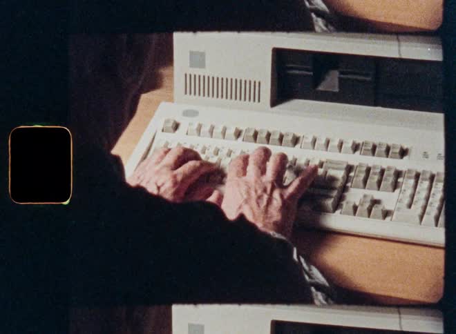 A close-up over-the-shoulder shot on film of an elderly person's hands typing on a vintage computer