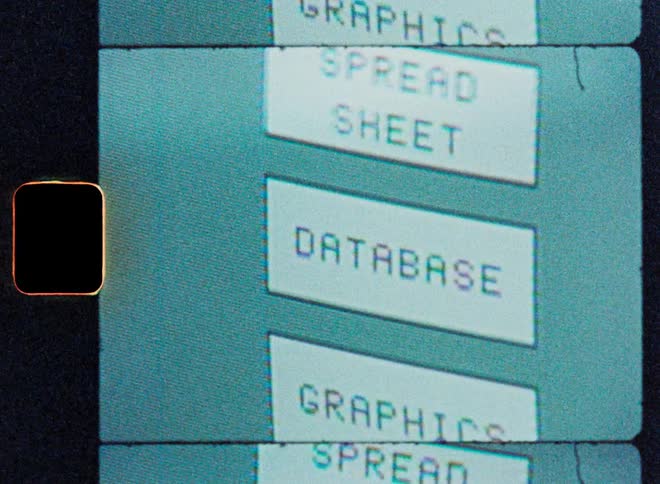 Vintage film scan captures a close-up of a retro computer monitor scrolling through a menu of early software applications