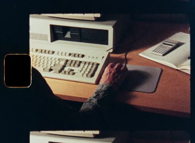 An over-the-shoulder view of a person typing on a vintage desktop computer, scanned from old 16mm film