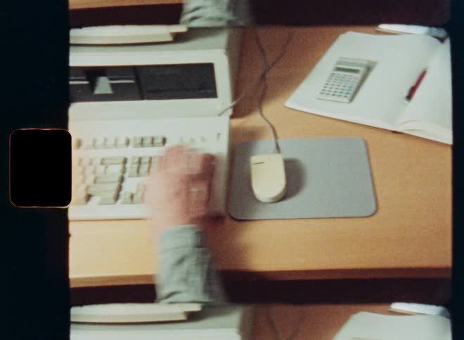 A high-angle vintage film shot of a person's hands typing on a retro personal computer keyboard from the 1980s or 1990s
