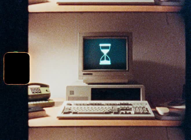 A vintage computer from the 1980s displays a loading hourglass icon on its screen in a grainy, retro-style film clip