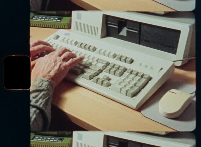 Archival film footage shows the hands of an older person typing on a vintage 1980s personal computer