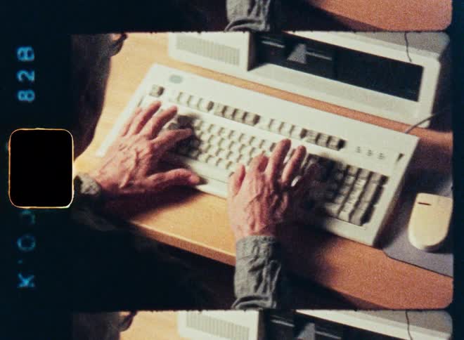 An over-the-shoulder shot of an elderly man's hands typing on a vintage computer keyboard and using a mouse, captured on grainy film