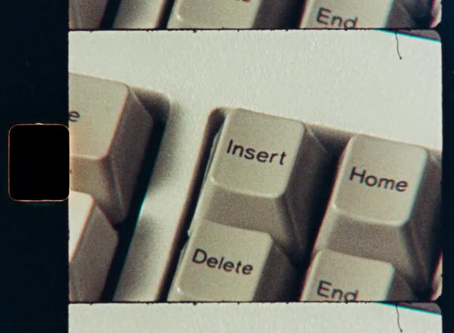 A vintage film scan shows a close-up of a finger pressing the delete key on an old computer keyboard