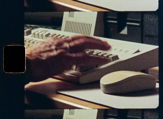 Vintage 16mm film footage shows a close-up of an elderly person's hands using a retro computer keyboard and mouse