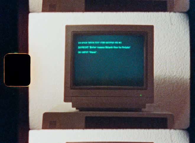 Close-up of a vintage computer monitor from the 1980s showing BASIC programming code being typed, with an authentic film look