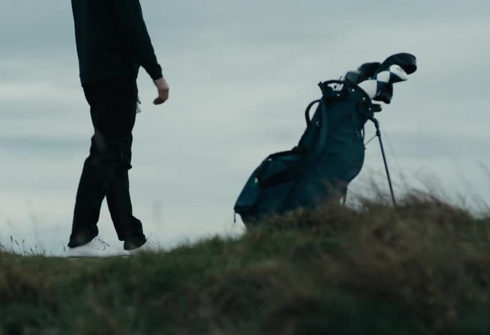 A golfer walks in slow motion across a grassy hill, setting down his golf bag on an overcast day
