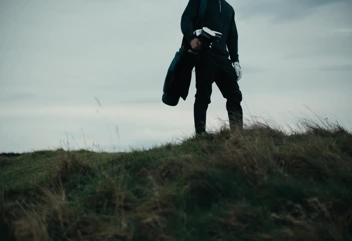 A golfer walks up a grassy hill in slow motion, carrying his bag of clubs before stopping to stand at the top