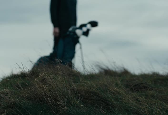 Low-angle shot of wild grass blowing in the wind as an out-of-focus golfer places their bag on a hill