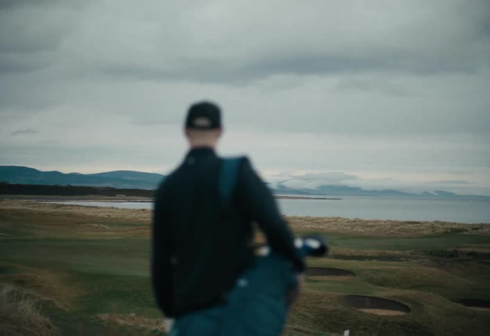 A golfer walks away from the camera, overlooking a vast seaside links golf course on an overcast day