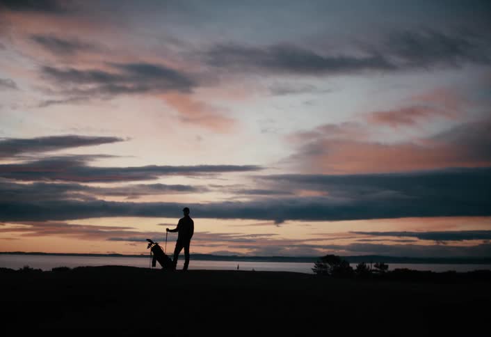A lone golfer is silhouetted against a beautiful pastel sky as they stand on a coastal course at sunset