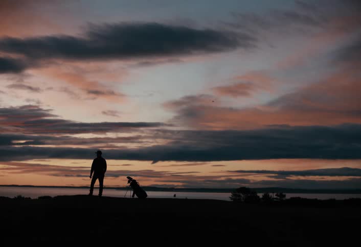 A golfer's silhouette is cast against a beautiful sunset as they take a swing on a coastal course