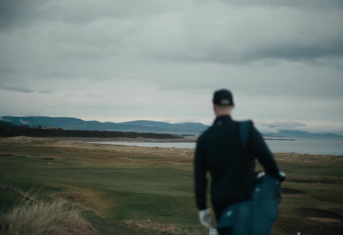 A golfer in slow motion carries his bag and walks onto a vast seaside links golf course on a dramatic cloudy day