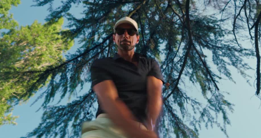 A man in sunglasses and a hat takes a powerful golf swing in slow motion, filmed from a low angle against the trees and sky