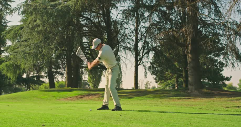 A man in a white hat and sunglasses takes a powerful swing with a golf club on a lush green course in slow motion