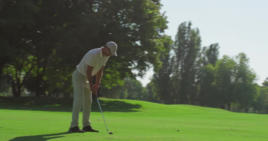 A man in a white polo and cap lines up his shot and swings his golf club in slow motion on a beautiful green course