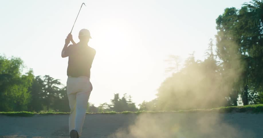 Slow motion shot of a professional golfer hitting a ball out of a sand bunker, with sand dust filling the air in the bright sunlight