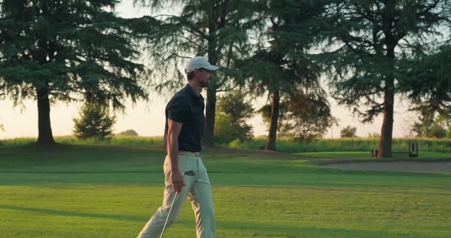 A man swings his golf club as his fluffy white Samoyed dog playfully chases after the ball across the green course at sunset
