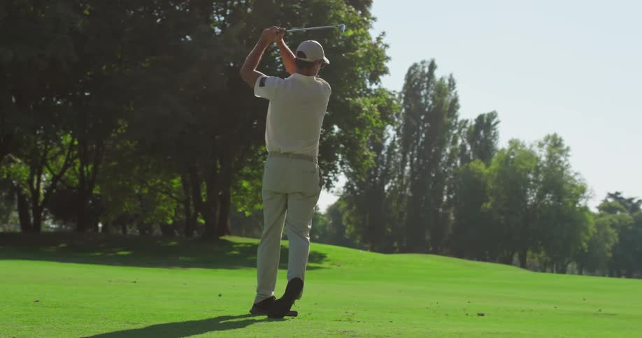 A male golfer executes a powerful swing on a sunlit, green golf course in slow motion