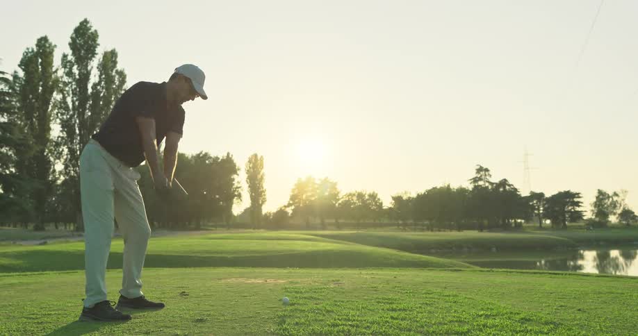 A male golfer in a white hat hits a powerful drive in slow motion on a beautiful course at sunset