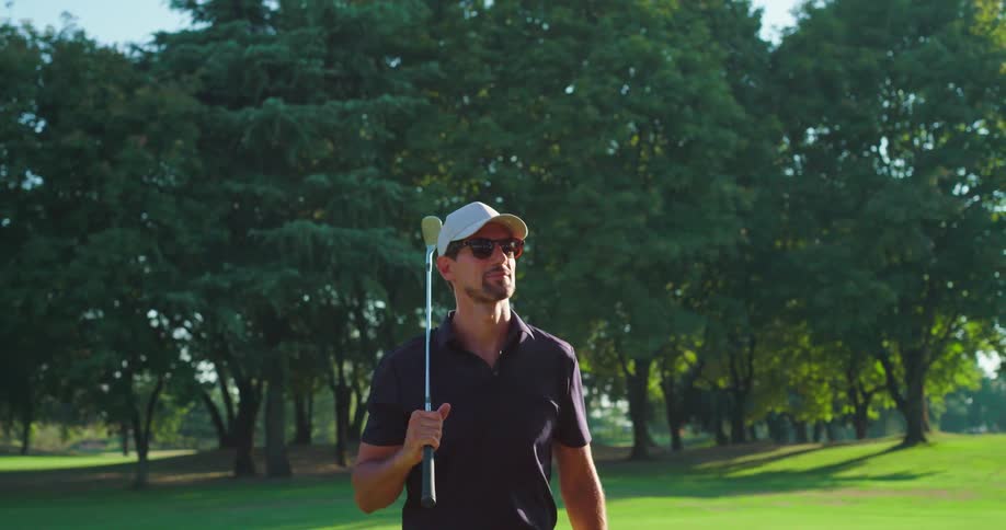 A handsome golfer in a cap and sunglasses walks confidently across a lush green course, holding his club on a sunny day in slow motion