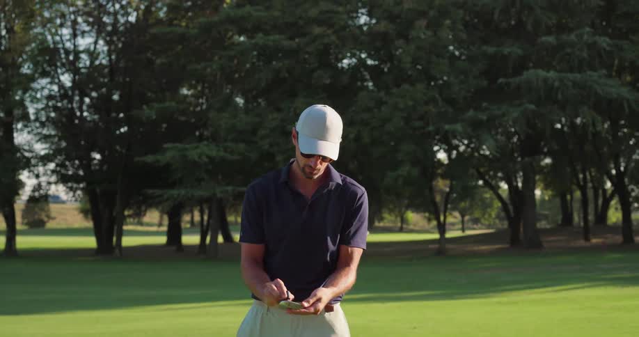 A male golfer in a cap and sunglasses swings his club on a green course and then cleans it while walking