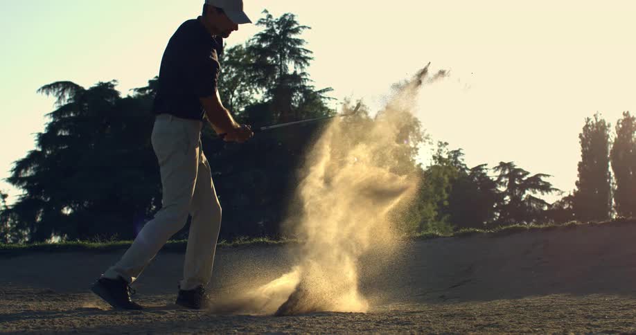 A silhouetted golfer hits a ball out of a sand bunker in dramatic slow motion as the sun sets