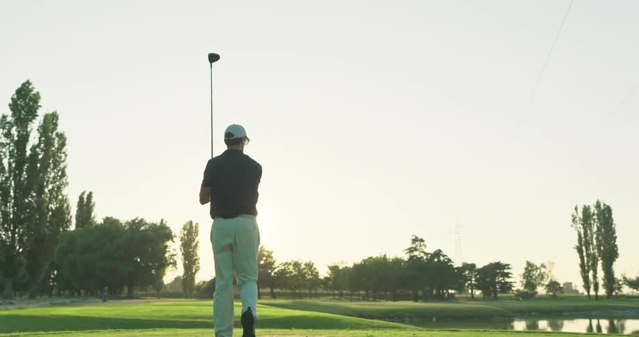 A male golfer takes a powerful swing in slow motion on a beautiful golf course during a golden sunset
