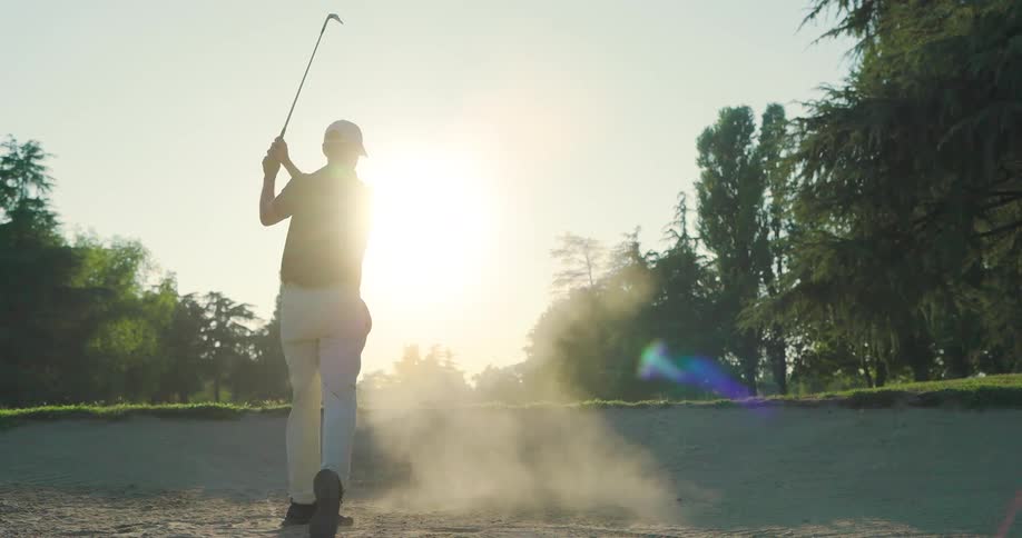A skilled golfer makes a powerful shot out of a sand bunker in slow motion, with the sun setting beautifully in the background