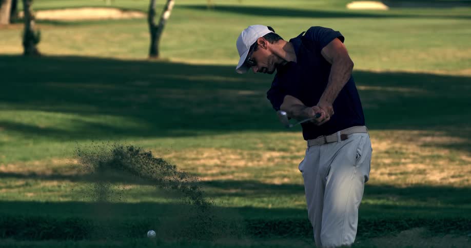A male golfer in a white hat hits a golf ball out of a sand bunker in super slow motion