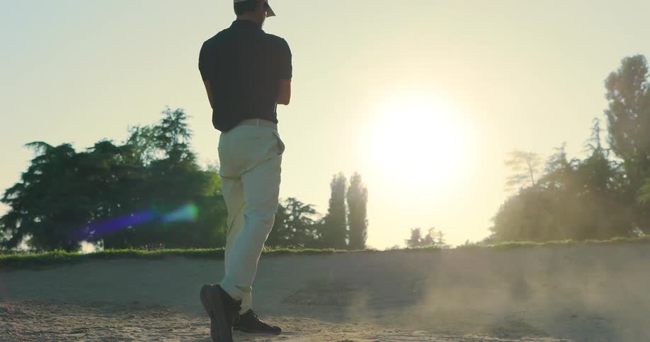 A golfer takes a powerful swing in slow motion, sending the ball and a cloud of sand flying from a bunker at sunset