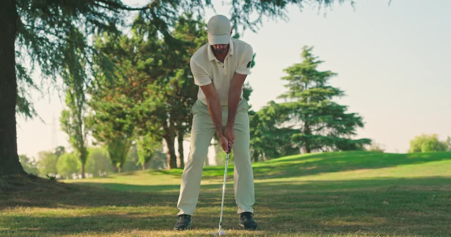 A male golfer in a white polo shirt and cap approaches his ball and hits a powerful swing in slow motion on a beautiful, sunny golf course