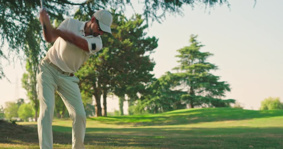 A male golfer in a white polo shirt takes a powerful swing at the ball on a sunny green course in slow motion
