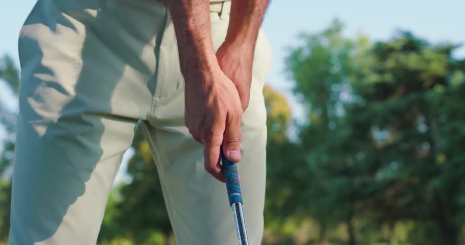 A close-up slow-motion shot shows a male golfer carefully adjusting his grip on a golf club on a sunny day