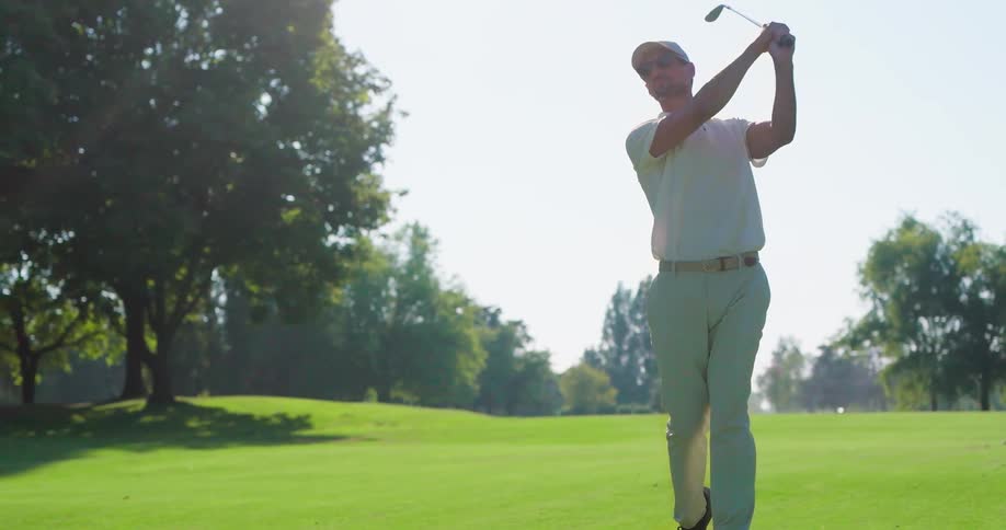 A male golfer executes a powerful swing in slow motion, sending the ball flying across the sunny, tree-lined fairway