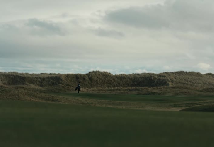 A lone golfer walks across a vast and empty links course with their bag under a moody, overcast sky