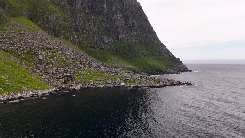 Static aerial view of a remote cabin nestled at the base of a towering green cliff along a rugged, rocky coastline