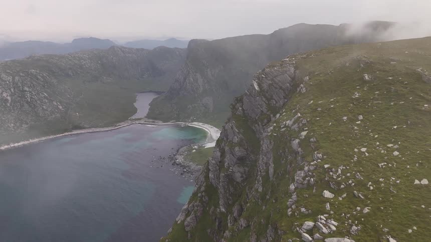 An aerial drone shot flies along a foggy, moss-covered cliff revealing a hidden bay with a white sand beach in a dramatic mountain landscape