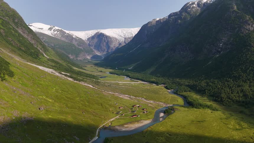 A stunning static aerial view of a remote village nestled in a lush green valley with a river, leading to a massive glacier in the mountains