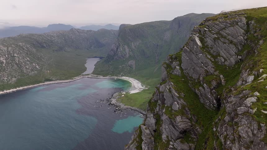 A majestic aerial view slowly flies along a dramatic green cliffside, revealing a secluded bay with a white sand beach and turquoise water nestled between mountains