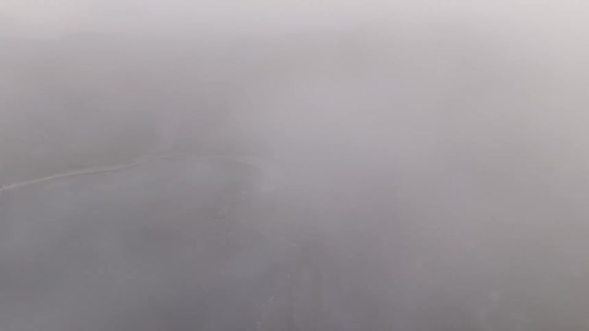 An aerial view descends through thick fog to reveal a stunning secluded beach with turquoise water nestled among dramatic green mountains