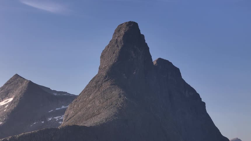 The majestic and rugged peak of Stetind mountain in Norway stands tall against a clear blue sky as tiny hikers reach the summit