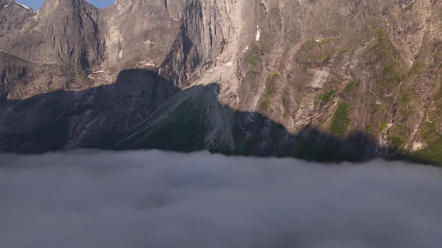An awe-inspiring aerial view tilts up from a thick layer of clouds to reveal a majestic, sunlit mountain range with jagged peaks against a clear blue sky