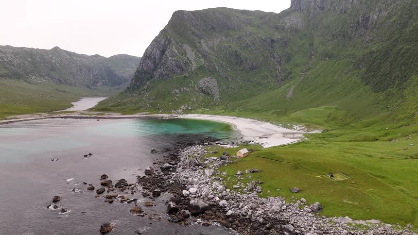 An aerial view slowly advances over a secluded bay with a white sand beach, turquoise water, and a lone cabin surrounded by lush green mountains in the Lofoten Islands, Norway